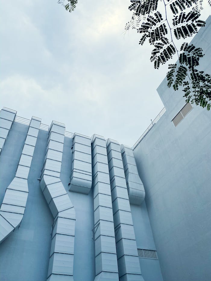 Pic 12 Low-angle view of industrial ductwork on a modern building exterior with sky and tree leaves.